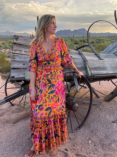 Woman in a vibrant floral dress standing next to a vintage wagon in a desert landscape, embodying an adventurous spirit and artistic background.